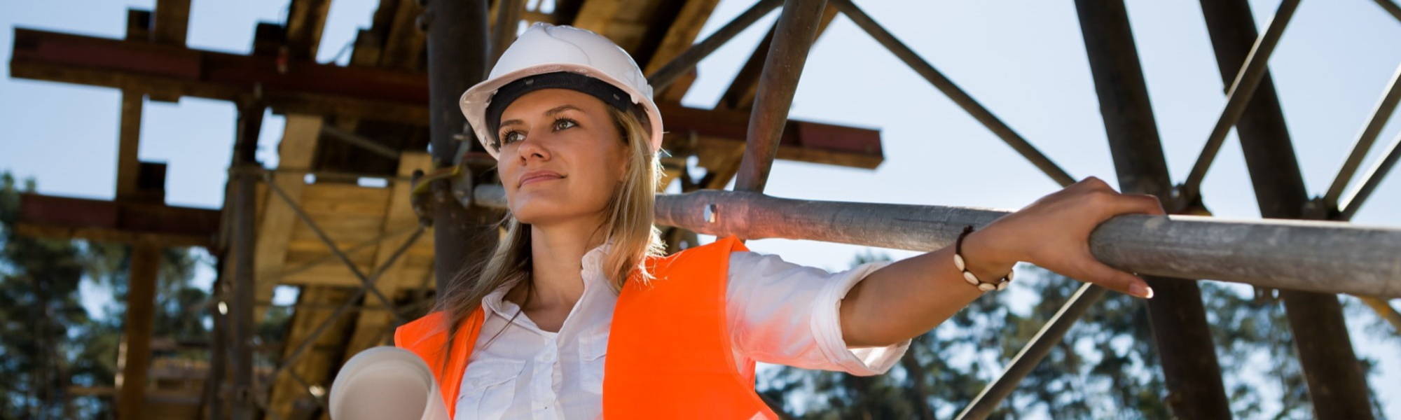 a woman standing next to scaffolding wearing a white hard hat and orange safety vest