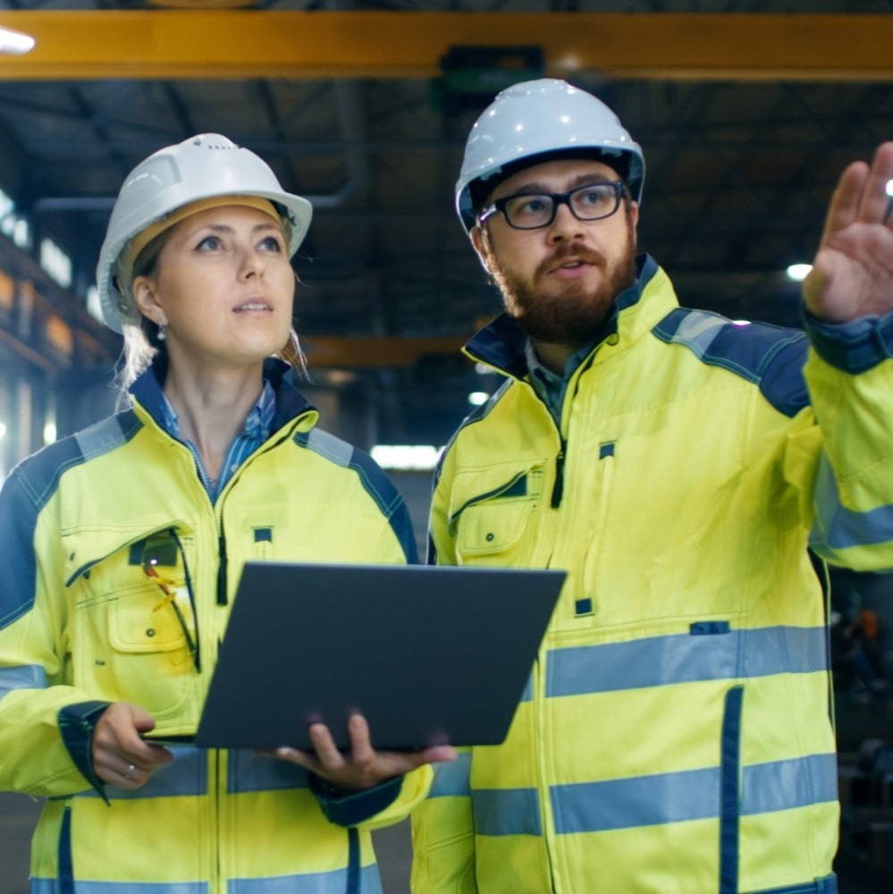 a man and a woman wearing PPE talking and holding a laptop