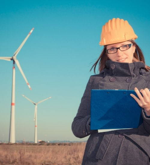 Lady wearing a yellow hard hat stood in front of wind turbines with a clipboard 