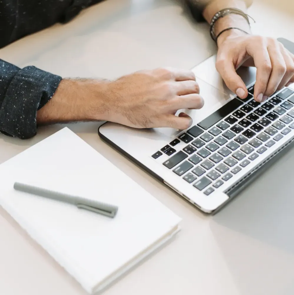 overhead view of a person's hands typing on a laptop with a notepad and pen