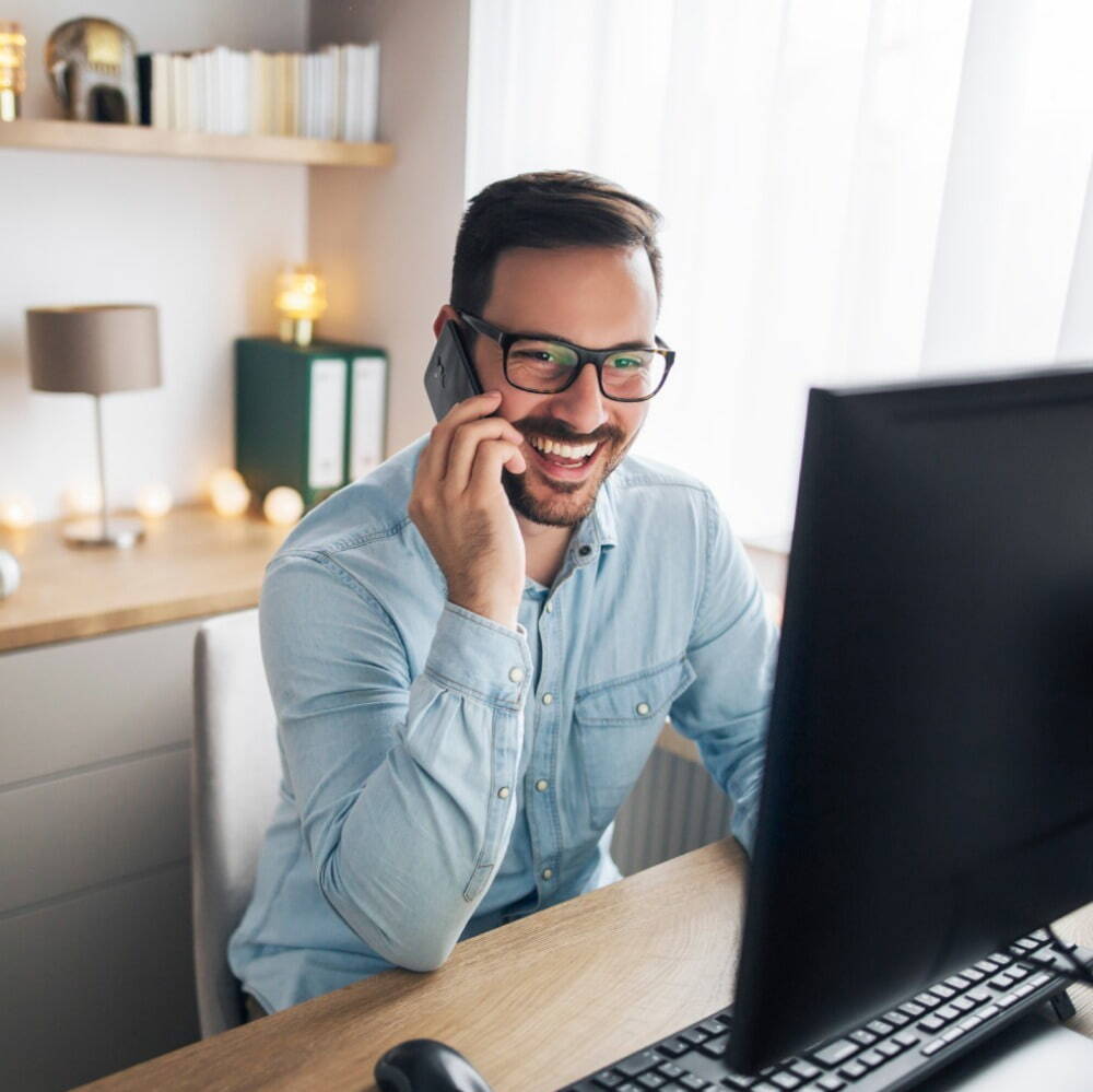 man talking on his phone looking at a computer, smiling 