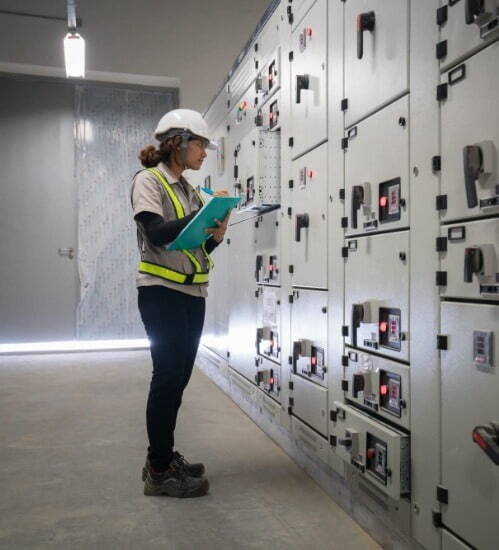 woman wearing a white hard hat checking equipment, holding a clipboard 