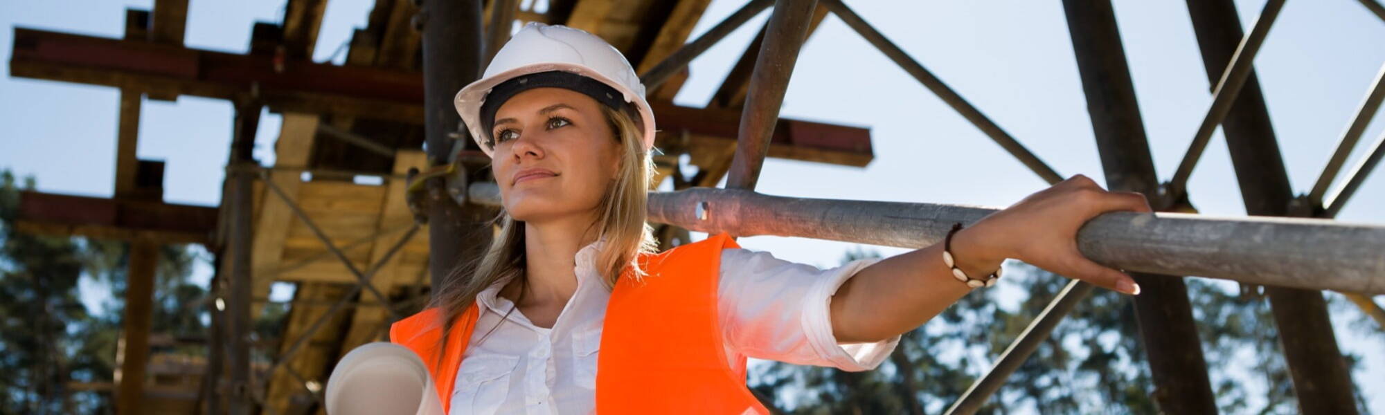 a woman standing next to scaffolding wearing a white hard hat and orange safety vest