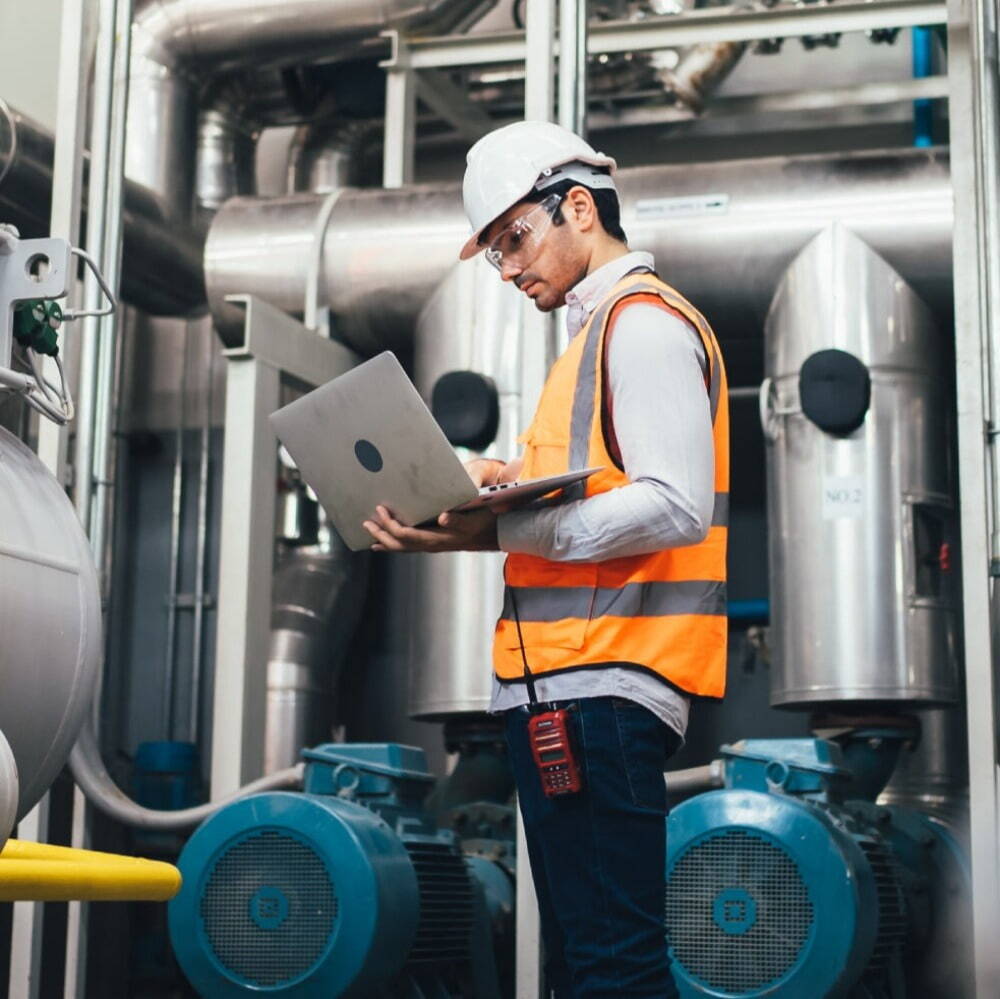 man with white hard hat and orange saftey vest stood up looking at a laptop that he's holding