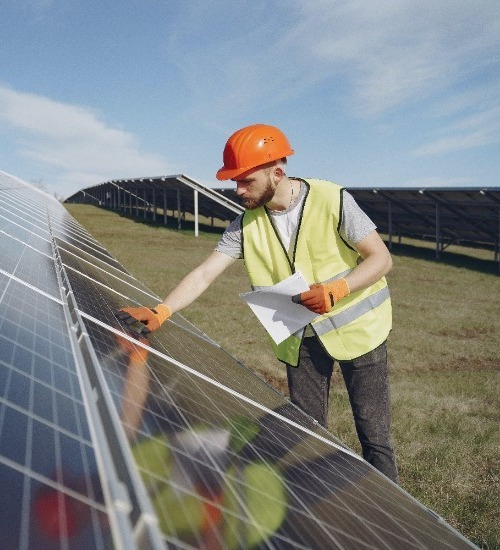 man looking at a row of solar panels wearing an orange hard hard and yellow safety vest