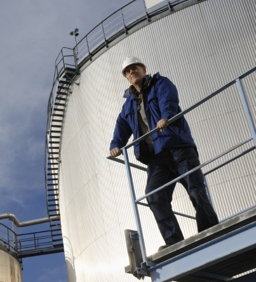 Man stood on balcony over looking the area with a white hard had and blue overalls 